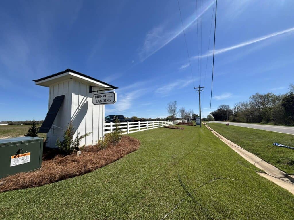 Landscaped entrance to Bienville Landing with a white fence, lawn, and ornamental plantings