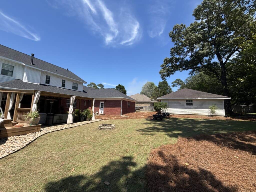 Residential backyard lawn with a covered patio, stone edging, and mature trees